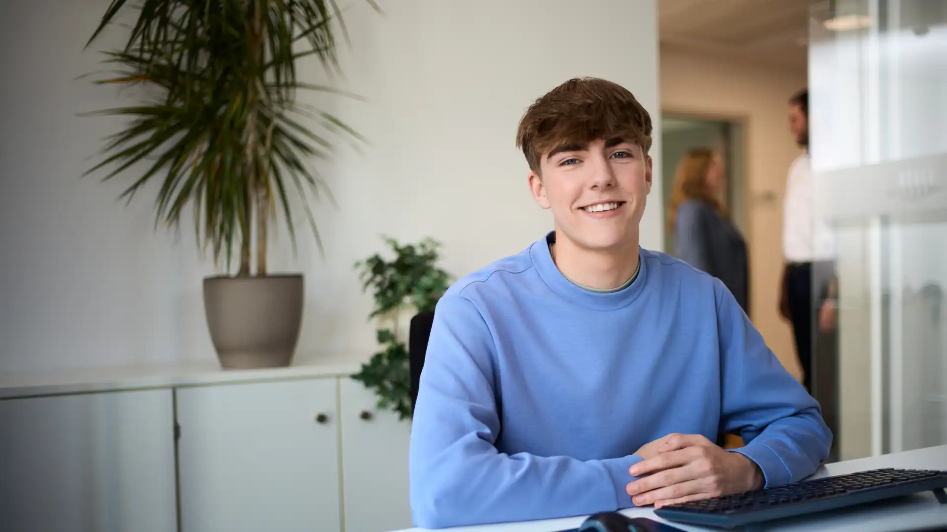 Ein Mann im blauen Sweatshirt sitzt an einem Schreibtisch und lächelt