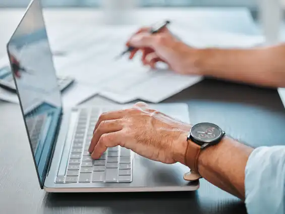 Eine Hand, die auf der Tastatur eines Laptops liegt, im Hintergrund eine andere Hand, die etwas notiert 